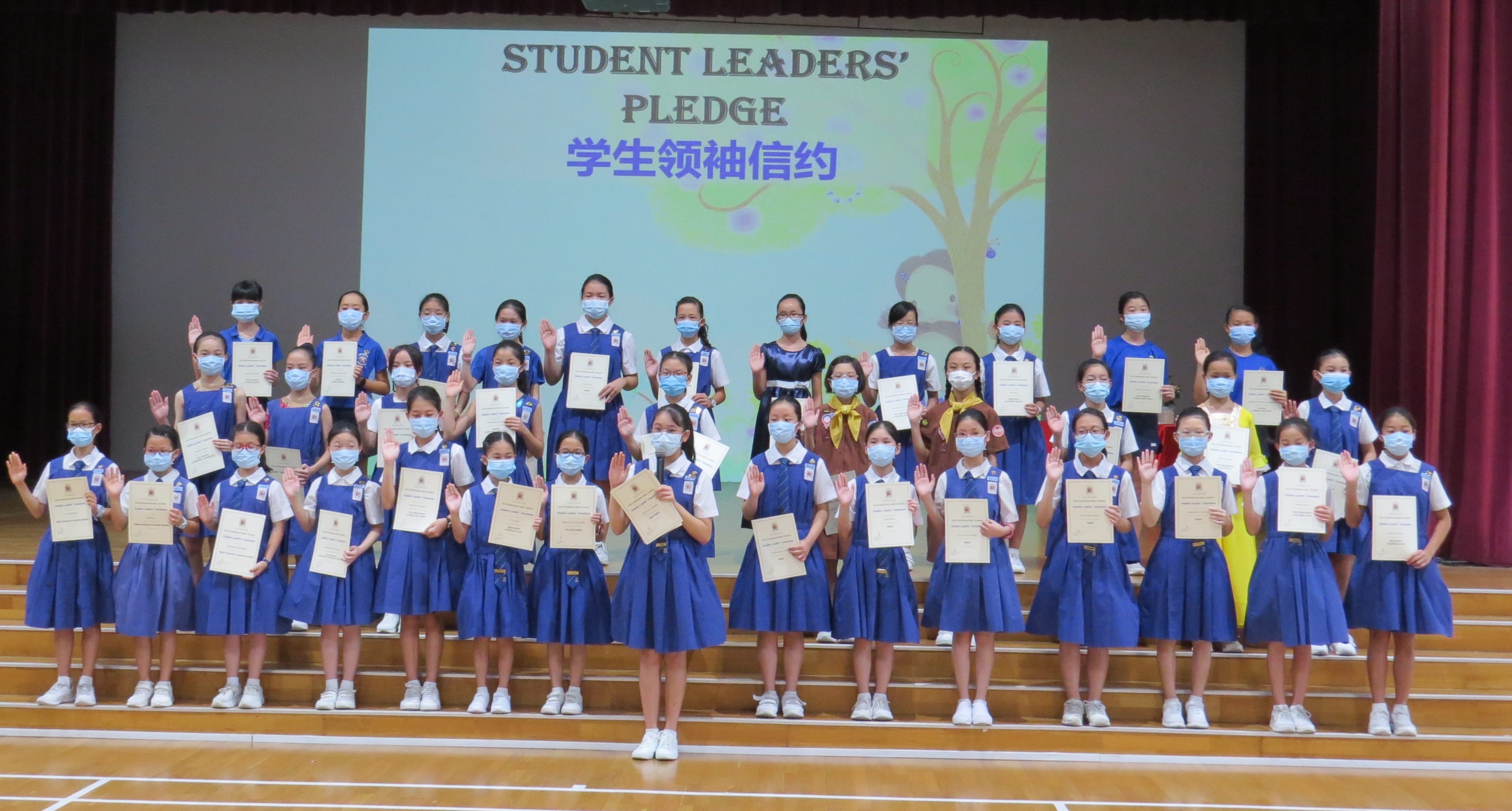 Students in blue uniforms and masks hold certificates and raise hands at a Student Leaders' Pledge ceremony on stage.